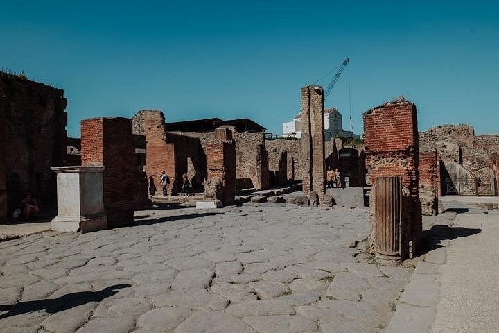 A basalt-paved street at Pompeii between high kerbs, excavation cranes beyond